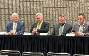 Four men sitting at a conference table in the front ready to speak to a group all with suit jackets on